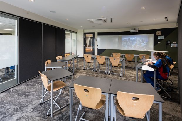 A modern classroom or training room with rows of movable tables and light timber chairs, dark acoustic wall panels, patterned carpet, large glazed partitions, recessed lighting, and a front projection screen. A few people are seated at the desks along the right side.