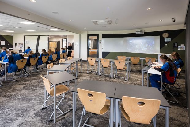 Modern flexible classroom or training room with rows of light timber student chairs and individual tables, a large projection screen on a dark feature wall, and glazed partitions opening to adjacent learning spaces. The room has grey patterned carpet, white ceilings with recessed lighting and exposed air-conditioning vents, and a bright, functional educational interior.
