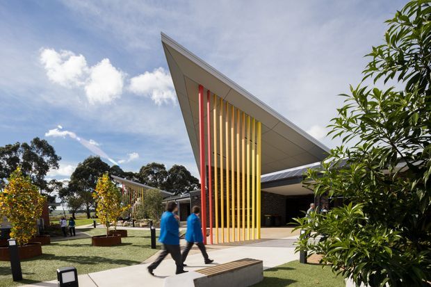 Exterior view of a contemporary public or commercial building with a dramatic sharply angled roof canopy and a tall facade screen composed of vertical multicoloured fins, including red, yellow, and timber-toned elements. The landscaped forecourt includes paved paths, raised planters, trees, and built-in concrete seating. People are walking through the space.