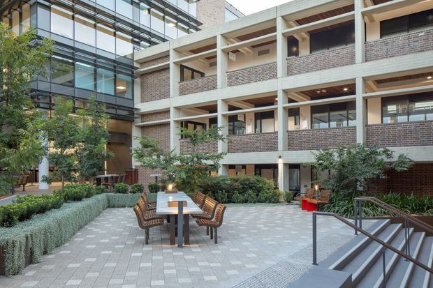 A landscaped urban courtyard adjoining a multi-storey contemporary brick and concrete building. The paved plaza includes an outdoor dining table with chairs, raised garden beds, shrubs and small trees, with stairs and metal handrails in the foreground. Large glazed sections and open recessed balconies create a sheltered communal outdoor space.