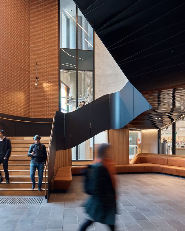 Interior view of a contemporary public or commercial atrium with a dramatic sculptural black stair and balustrade sweeping diagonally through the space. The lobby features exposed red brick walls, tall glazing, warm timber wall panels, built-in tan upholstered bench seating, dark tiled flooring, and linear architectural lighting. People are present, with motion blur in the foreground. A retail or service area is visible beyond the lobby.