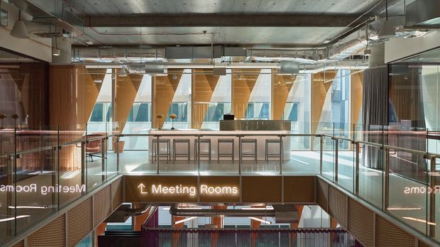 A bright contemporary commercial interior showing an elevated meeting-room and breakout area around a central double-height void. Glass balustrades, exposed concrete and services, extensive ductwork, timber-lined walls, and large angular glazed facade elements create an industrial-modern workplace atmosphere. Illuminated signage identifies meeting rooms, with a stair or lower-level circulation area visible below.