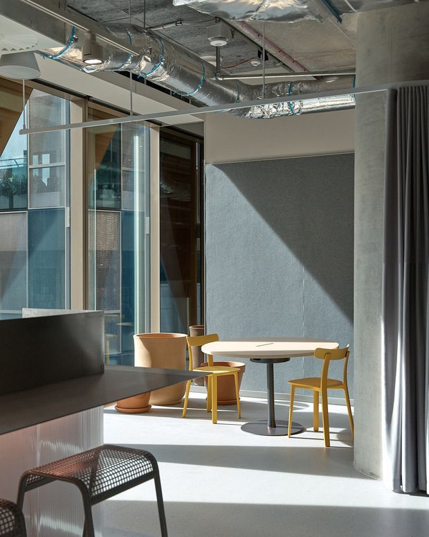 Contemporary commercial interior with a small dining or breakout area beside a dark counter. The space features exposed silver HVAC ductwork and services, concrete columns, full-height glazing, a grey acoustic or textured wall panel, pale flooring, a round light timber table, and mustard-yellow chairs. Strong sunlight creates a diagonal shadow across the wall and floor, while dark curtains frame the right side.