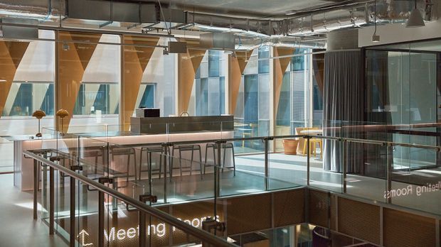 A contemporary multi-level commercial interior with an open mezzanine or stair landing, glass balustrades, exposed ductwork and services, suspended pendant lights, timber-lined structural elements, and extensive floor-to-ceiling glazing. A long communal counter with stools overlooks the lower level, while a glazed meeting room is visible to the right. Reflected or reversed lettering appears on the glass.