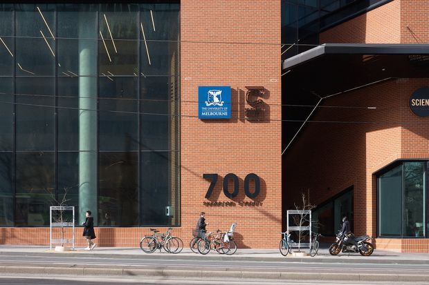 Exterior street-level view of a contemporary university or science building. The facade combines extensive dark glazed curtain walls with red brick masonry, large metal numerals reading 700, and a University of Melbourne sign. A recessed entrance or passage sits to the right beneath a projecting dark canopy, with bicycles and pedestrians along the sidewalk.