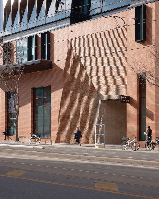 Street-level view of a contemporary multi-storey brick building with a highly articulated geometric brick facade. The elevation combines warm red brickwork, deep-set vertical windows, dark metal framing, reflective upper-level glazing, and a prominent stepped brick canopy or entry feature. A sign identifies a creative or cultural space, while bicycles, pedestrians, and a tram or roadway occupy the foreground.