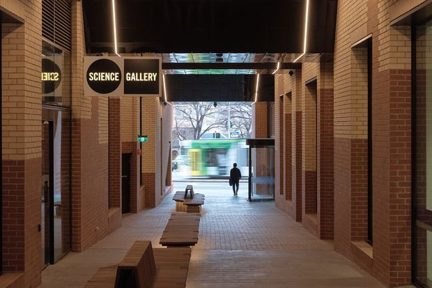 A covered urban passageway leading toward a science gallery entrance, framed by warm red-brown brick walls and brick columns. The corridor has paved flooring, integrated timber bench seating, dark overhead ceiling panels with linear strip lighting, exposed skylight glazing, signage, and glazed storefront or gallery openings. A blurred city street and tram are visible beyond the passage.