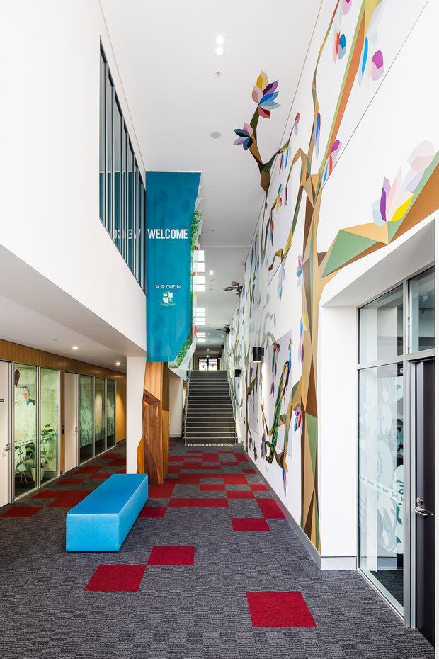 A bright, contemporary institutional or commercial hallway with a tall white interior volume, a large colourful abstract mural, patterned grey and red carpet tiles, extensive glazed partitions, a vivid blue upholstered bench, and a staircase leading upward. A turquoise welcome banner hangs in the corridor, reinforcing the public or educational setting.