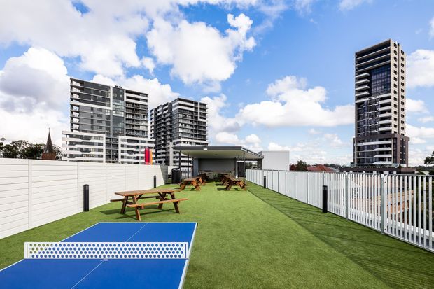 Outdoor communal recreation area with artificial turf, picnic tables, a covered pavilion, and a blue recreational court in the foreground, enclosed by white fencing. Several modern high-rise apartment buildings are visible beyond the site.