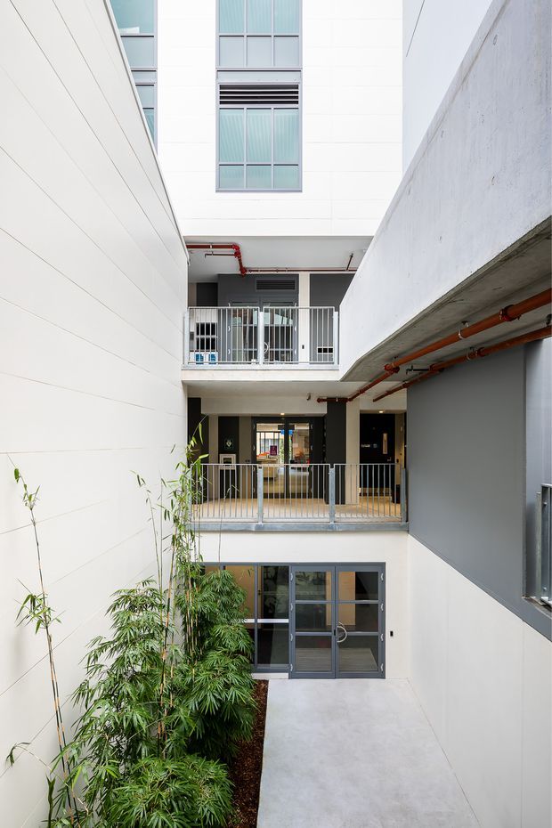 A bright multi-level interior courtyard or atrium within a contemporary building. White horizontal wall cladding, exposed concrete balconies and soffits, grey metal-framed glazing and railings, and visible red service pipes create a clean institutional or commercial character. Bamboo planting lines the lower walkway, with several levels of balconies and circulation spaces overlooking the courtyard.