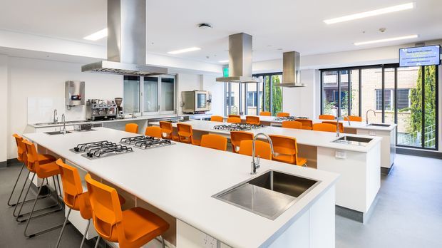 Bright, spacious commercial teaching kitchen with multiple large white island workbenches, stainless-steel sinks and cooktops, suspended stainless-steel extraction hoods, and rows of orange bar stools. Extensive glazing provides daylight, with visible appliances, service areas, and a wall-mounted display screen.