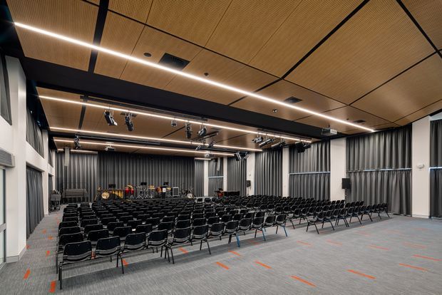 A large modern auditorium or performance and events room viewed from the rear, with many rows of black chairs facing a stage area. The room features stage lighting and audio equipment, dark acoustic curtains, a wood-toned slatted acoustic ceiling with black framing, grey carpet flooring with orange accent markings, and large wall-mounted acoustic panels.