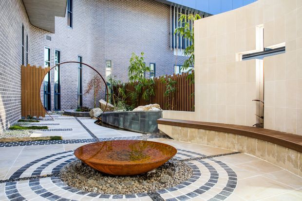 A contemporary landscaped courtyard with patterned circular paving, a rusted metal water bowl feature set in pebbles, a tiled rendered wall with a built-in timber bench, raised planting beds, timber fencing, and surrounding light brick walls. The courtyard includes a curved metal garden arch and a small water feature or pond.