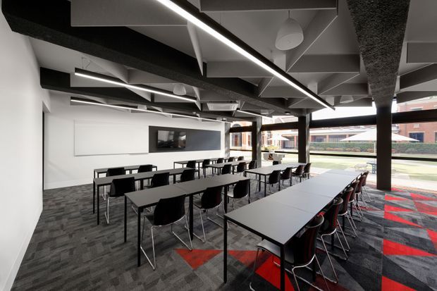 Open-plan contemporary commercial interior arranged as a flexible meeting, training, or breakout space. Multiple long rectangular tables with black chairs occupy a carpeted room, with a wall-mounted display at the far end. The room features exposed structural and services elements across the ceiling, linear integrated lighting, dark columns, and large glazed openings overlooking an urban exterior. The palette is predominantly white, charcoal, black, and grey with bold red carpet accents.