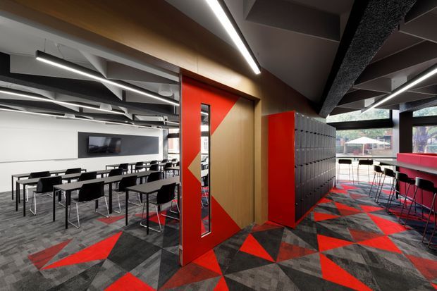 Modern commercial interior featuring a flexible dining or breakout area with rows of black tables and chairs, a red high counter with stools, and a bold geometric carpet in charcoal grey and red. A red and timber pivot door with a narrow glazed panel opens toward the seating area, while grey locker-style cabinetry and a large wall-mounted screen are visible. The ceiling has exposed services, acoustic elements, timber soffit panels, and linear LED lighting, creating a contemporary industrial workplace or hospitality setting.