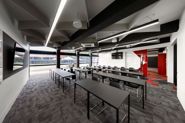 A contemporary commercial interior with rows of black rectangular tables and chairs arranged in a large open meeting, training, or breakout space. The room features dark patterned carpet, white walls, exposed black ceiling beams and services, linear LED lighting, wall-mounted screens, and large windows overlooking an exterior sports facility. A bold red-and-black accented corridor or room is visible at the rear.