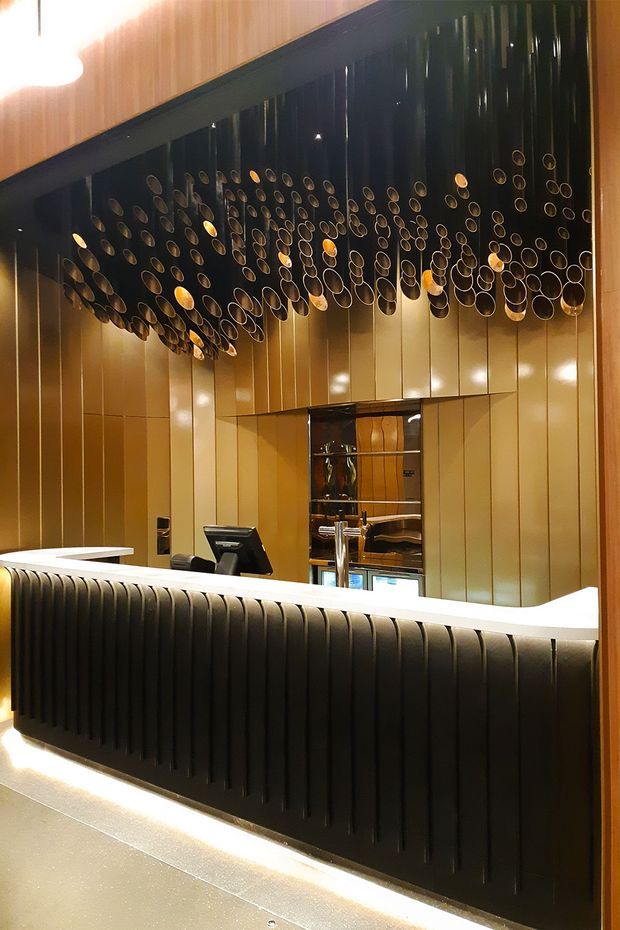 A dramatic commercial reception or hotel front desk featuring a long curved counter with a glossy white top and deeply fluted dark front. Behind it, reflective gold-toned vertical wall panels and a large sculptural installation of suspended circular metal elements create a luxurious, theatrical interior. A small service opening and point-of-sale equipment are visible behind the desk.