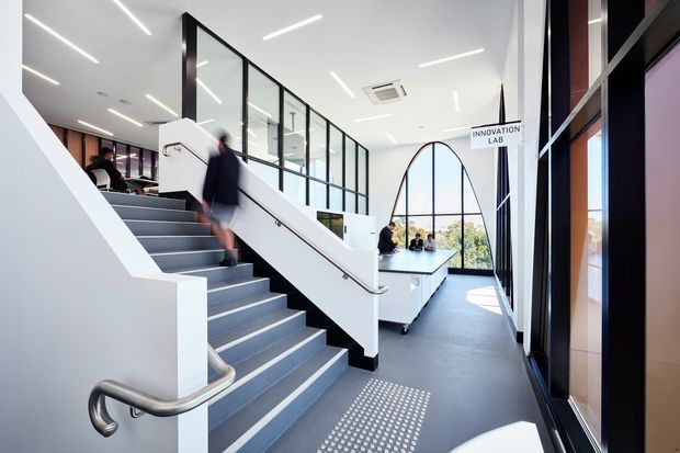 Bright contemporary commercial interior showing a stairway and upper-level landing, with a glass-partitioned innovation lab, white balustrades, dark flooring, linear ceiling lights, and large arched glazing. People are present in the background, and an accessible tactile floor strip is visible near the stairs.