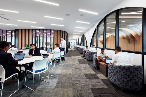 Bright contemporary commercial workspace with rows of shared desks, computer workstations, task seating, patterned lounge chairs, and a carpeted circulation zone. A striking curved glazed partition runs along the right side, revealing warm timber curved ceiling or wall detailing beyond. The space has linear recessed lighting, dark-framed glazing, and an open collaborative office layout.