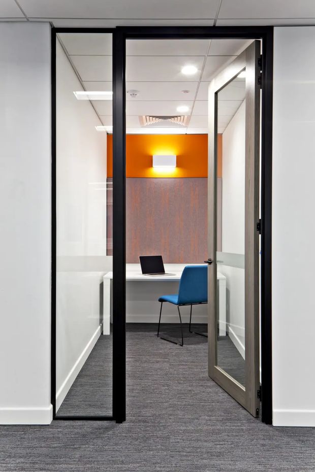 A compact enclosed office or meeting room viewed from a corridor through a black-framed glazed door and side panel. The room has a white desk with a laptop, a single blue office chair, dark grey carpet, white walls, a wood-toned feature wall, and a vivid orange accent wall with a wall light. The space has a suspended acoustic tile ceiling with recessed lighting and visible air-conditioning ventilation. The open door and glass partitions create a contemporary commercial office setting.