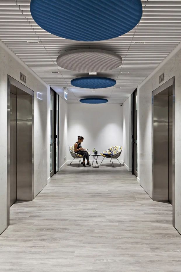 A modern commercial elevator lobby or hallway with two stainless-steel elevator doors, pale grey wood-look flooring, light textured walls, and a white linear slatted ceiling. Large circular acoustic ceiling panels in navy blue and grey create bold overhead accents. At the far end, two patterned lounge chairs and a small round table form a compact waiting area.