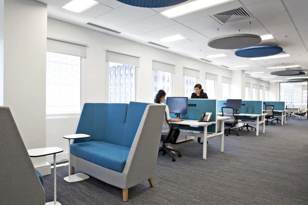 Bright contemporary open-plan commercial office with rows of workstations, computers, ergonomic office chairs, turquoise desk screens, and a blue-and-grey high-backed lounge booth in the foreground. The space has extensive windows, grey carpet flooring, white walls, a suspended acoustic tile ceiling with recessed lighting, linear air-conditioning grilles, and multiple circular blue and grey acoustic ceiling panels.