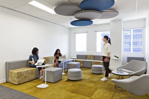 Bright contemporary office lounge and waiting area with modular grey upholstered seating, mustard patterned bench cushions, ottomans, small side tables, and a large mustard-toned carpet. White walls, suspended linear lighting, large windows, and layered blue and grey acoustic ceiling baffles create a modern collaborative workspace atmosphere.