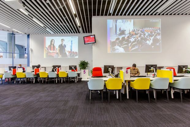 A bright contemporary commercial learning or collaboration space with rows of computer workstations, colourful red, yellow, white and grey chairs, large projection screens, and a linear slatted ceiling. The room has glazed partitions, grey carpet flooring, white walls, and an open, highly functional layout.