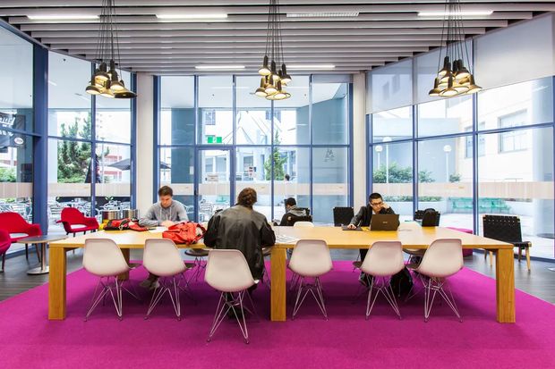 A bright contemporary open-plan workspace with a long communal timber table, multiple task chairs, people working on laptops, and a vivid magenta carpet. Floor-to-ceiling glazed walls provide views into an urban courtyard or neighbouring building. The ceiling features linear slatted detailing and suspended decorative pendant lights, while blue-framed glazing and transparent partitions define the office interior.