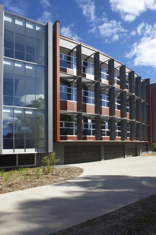 Exterior view of a contemporary multi-storey institutional or commercial building. The facade combines extensive floor-to-ceiling glazed curtain walling with dark vertical metal fins, reddish-brown panel cladding, white-framed windows, projecting sunshade canopies, and a recessed ground-level garage or service area. A paved driveway and minimal landscaped planting sit in front of the building under a bright blue sky.