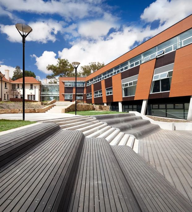 Exterior view of a contemporary institutional building arranged around a landscaped courtyard and stepped public seating area. The long red-orange brick facade features dark horizontal panels, extensive strip windows, projecting angled window bays, and a glazed connecting walkway. Broad dark timber-look decking and tiered concrete steps form an amphitheatre-like outdoor space, with modern black lamp posts and an older pitched-roof building visible nearby.