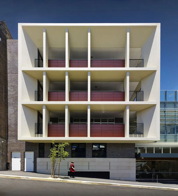 Front-facing exterior photograph of a contemporary multi-storey apartment building. The facade is organised into three stacked open balcony levels framed by deep white rectangular concrete projections and slender vertical columns, with dark metal balustrades and reddish timber-toned privacy screens. The lower level combines dark brickwork, pale wall panels, glazing and a recessed entry or commercial frontage. A neighbouring glazed structure is visible at right, with clear blue sky above.