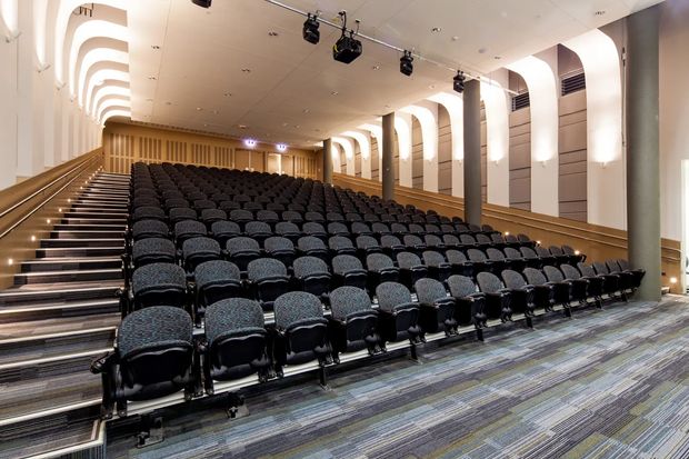 Interior view of a large tiered auditorium or lecture theatre with rows of dark upholstered seats, stepped aisles, acoustic wall panels, repeated arched wall details, warm wood finishes, carpeted flooring, and ceiling-mounted stage lighting.