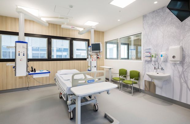 A bright, modern hospital patient room with a centrally positioned adjustable bed, overhead medical equipment, wall-mounted monitors, clinical cabinetry and services, a handwashing basin, and two green visitor chairs. The room has pale timber-look wall panelling, grey resilient flooring, white ceilings, large windows, and a patterned feature wall with integrated medical fixtures.