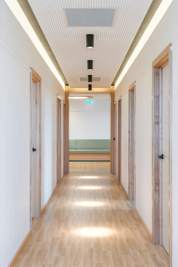 Bright, contemporary interior corridor with pale timber flooring, white walls, and multiple timber-framed doorways on both sides. The ceiling features a linear ribbed acoustic finish, recessed perimeter lighting, and black cylindrical downlights. A softly upholstered green bench is visible at the end of the hallway beneath an illuminated exit sign.