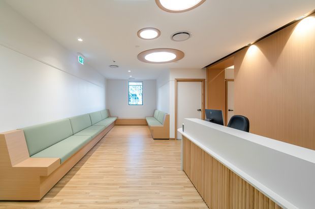 Bright contemporary reception area with a long pale green built-in bench along the left wall, a smaller matching seating area at the rear, and a timber-fronted reception desk with a white counter in the foreground. The space has light timber flooring, warm vertical timber wall panelling behind the desk, white walls, recessed and circular ceiling lights, and an accessible, uncluttered layout.