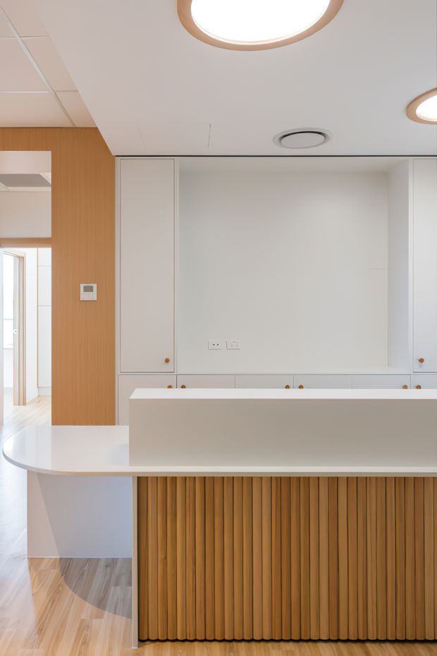 Bright contemporary kitchen interior featuring a large white island with a curved end and vertically fluted timber front, white integrated cabinetry, a warm timber-panelled wall, pale timber flooring, and a clean white ceiling with circular recessed lighting. A hallway or adjacent room is visible at left.