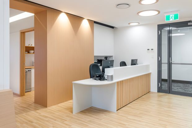 Bright contemporary commercial reception area with a curved white reception desk, vertical light-wood panelled front, black office chairs and computer stations. The space has pale timber flooring, a light-wood feature wall and doorway leading to a kitchenette or staff area, white walls, recessed circular ceiling lights, and glazed dark-framed double doors.