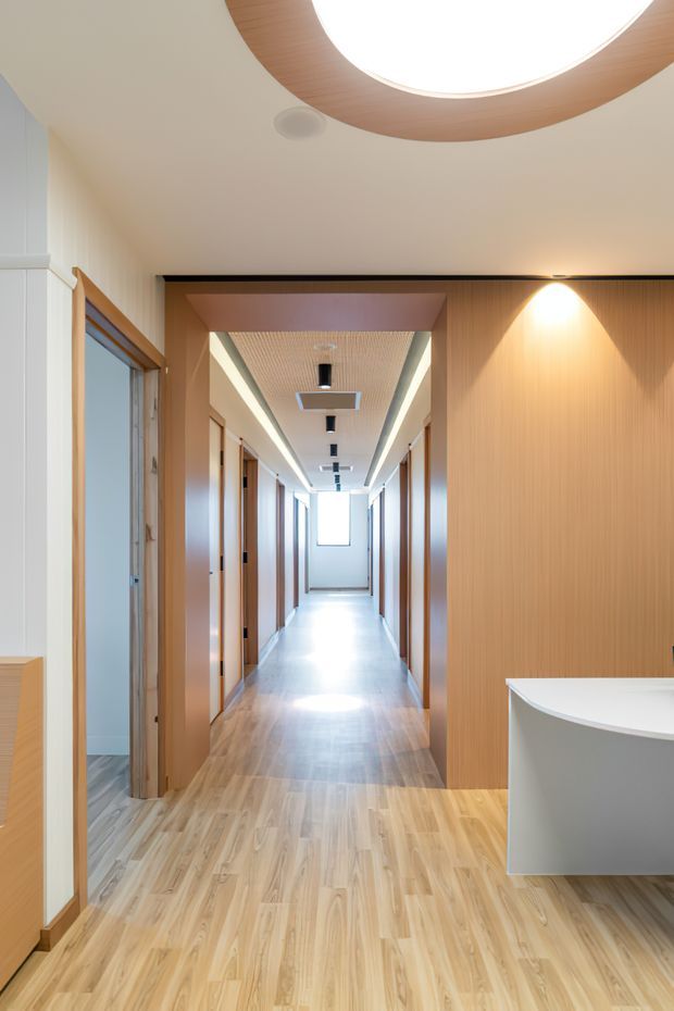 A bright, contemporary interior corridor viewed from an open vestibule or reception-like area. The long hallway has light timber flooring, timber-lined walls and door frames, recessed linear lighting, and multiple rooms opening along both sides. A curved white built-in counter or vanity is visible in the right foreground, while a large circular timber-trimmed ceiling light forms a distinctive feature overhead. The space is clean, warm, and highly ordered, with strong linear perspective and abundant daylight at the far end.