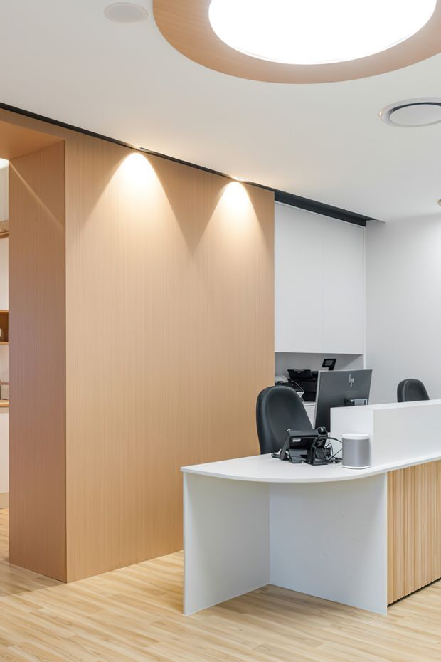 A clean, contemporary commercial reception area with a curved white reception desk, black office chairs, computer equipment, and a small cup. The space features pale timber wall panelling and matching timber flooring, white walls and ceiling, integrated warm spotlights, and a large circular ceiling light with a timber surround. A compact workstation is visible behind the reception counter.