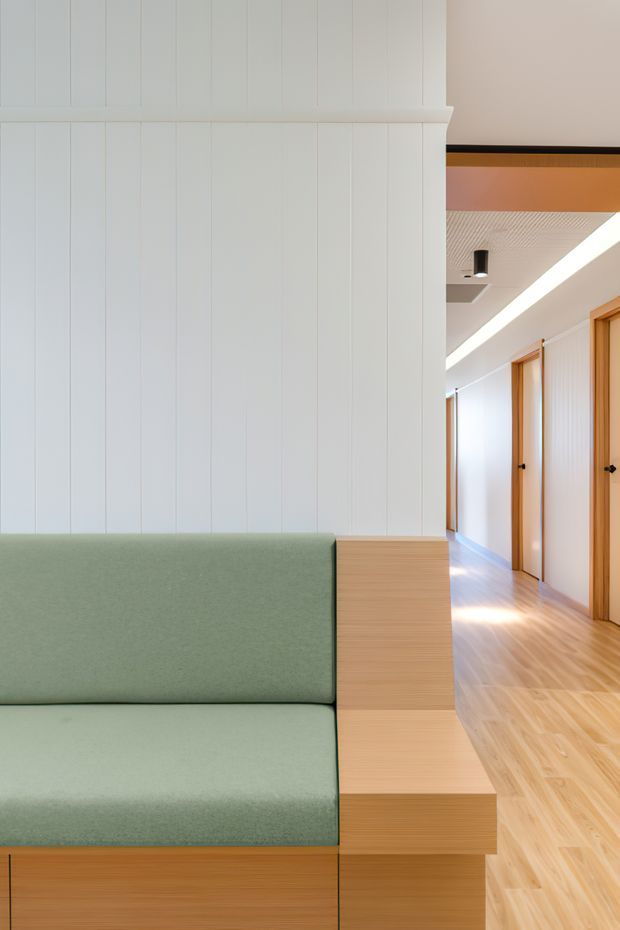 Bright contemporary interior showing a built-in pale green upholstered bench or sofa integrated with warm light timber cabinetry. White vertical wall panelling rises above the seating, while a timber-floored hallway with timber-framed doors extends into the background. The space features clean lines, concealed or minimal detailing, and linear ceiling lighting.