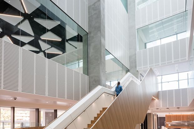 A bright, contemporary multi-level commercial interior with a prominent sculptural staircase and escalator-like circulation element. The space features extensive glass balustrades and partitions, pale timber-clad stair surfaces, perforated white acoustic wall and ceiling panels, exposed concrete, geometric pendant lighting, and a large open atrium with abundant daylight.