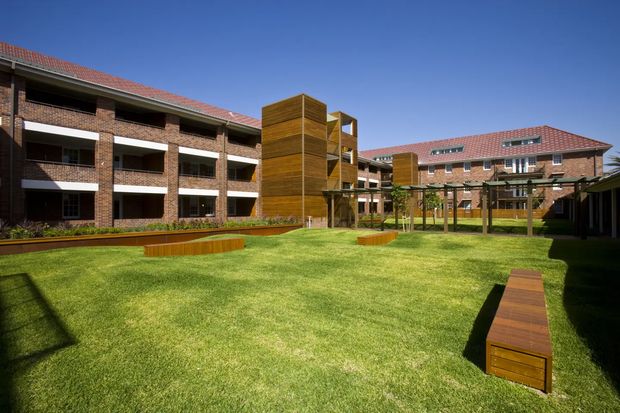 Exterior view of a multi-storey brick apartment or institutional complex arranged around a landscaped central courtyard. The buildings have red tiled hipped roofs, repetitive balcony bays, dark window openings, and prominent timber-clad vertical circulation or feature structures. The courtyard is predominantly lawn with long timber bench seating, raised planting beds, and a simple pergola-like walkway. Clear blue sky and strong sunlight create a bright, open setting.