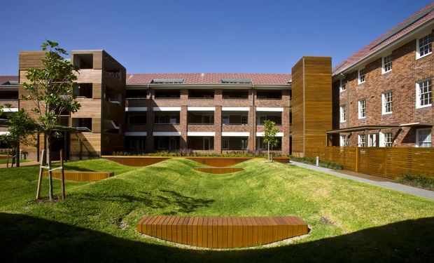 Exterior view of a multi-storey brick residential or institutional complex arranged around a landscaped courtyard. The buildings feature red tiled pitched roofs, white-framed windows, timber-clad vertical sections, balconies, and a gently mounded lawn with curved timber seating or edging.