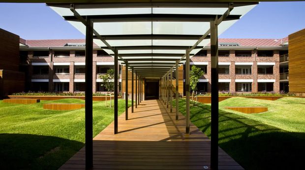A landscaped courtyard viewed along a straight timber boardwalk beneath a long open pergola with repeated dark structural posts and translucent roof panels. The path leads toward a covered entrance, with manicured lawns, raised circular garden beds, and a multi-storey red-brick building with regular windows and a low pitched metal roof surrounding the space.