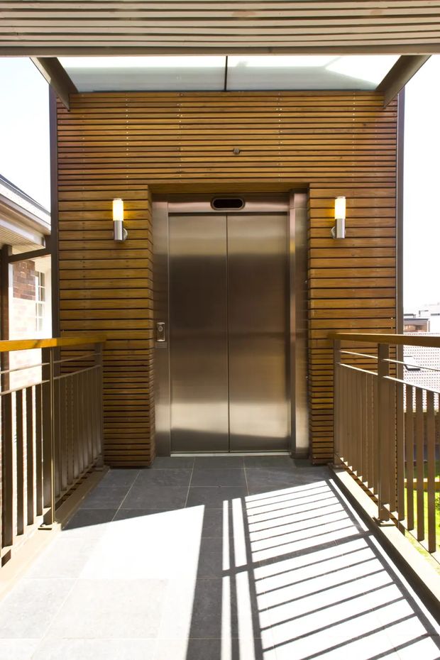 A bright, modern exterior lift entry on an upper-level walkway or balcony. The stainless-steel elevator doors are framed by warm horizontal timber battens, with slim wall sconces on either side and a translucent overhead canopy. Dark tiled flooring and metal balustrades surround the landing, with strong sunlight casting geometric railing shadows.