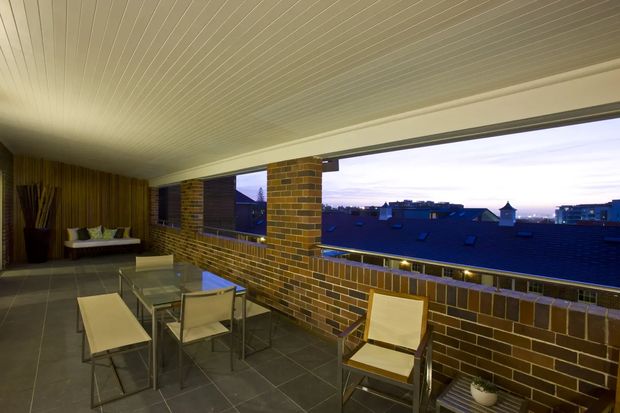 A covered rooftop terrace or balcony at dusk, furnished with a glass dining table, several light-coloured outdoor chairs, and a bench. The space has a dark tiled floor, exposed brown brick walls and balustrade, a timber-lined wall, and a low white slatted ceiling. Large open glazing frames provide views across neighbouring rooftops and the evening sky.