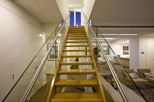 Interior view looking up a contemporary open-riser staircase with warm timber treads, stainless-steel handrails, and clear glass balustrades. Built-in white cabinetry lines the left side, while a partially visible living area with sofas and cushions is below and to the right. A bright landing and upper-level opening are visible at the top.