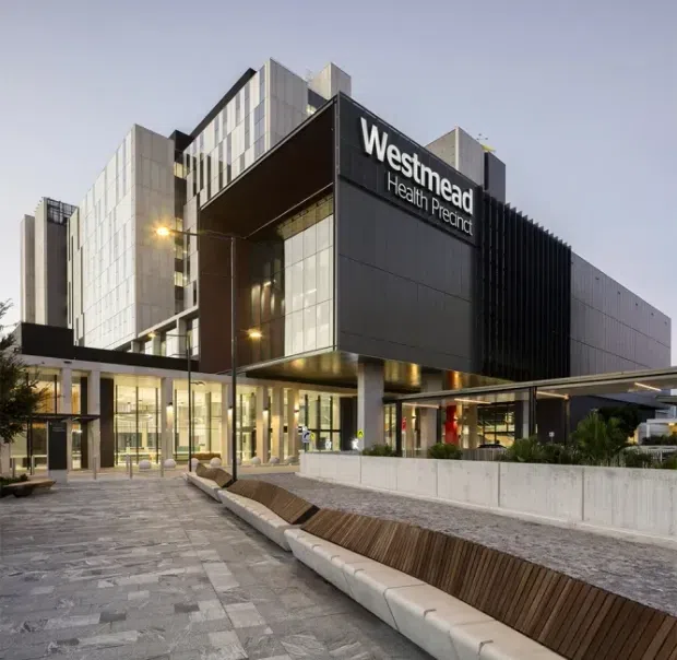 Exterior architectural photograph of the Westmead Health Precinct, a contemporary multi-storey healthcare building. The facade combines dark metal cladding, extensive glazed curtain walls, projecting box-like volumes, vertical fins, and a broad covered entrance canopy. A paved forecourt with integrated timber-and-concrete seating and landscaped planting occupies the foreground.