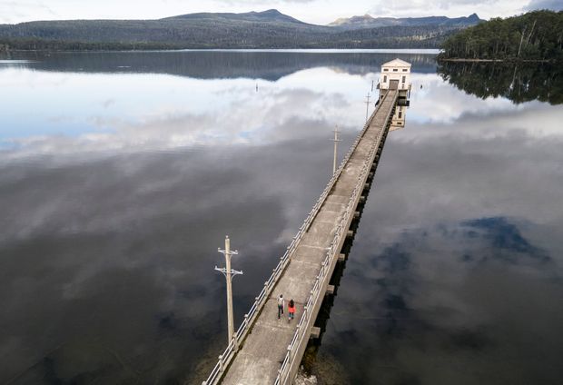 Aerial view of a long concrete pier or bridge extending across a calm mountain lake toward a small pale utility or pump house. Railings and regularly spaced poles line the structure, with forested mountains and reflected clouds surrounding the water.